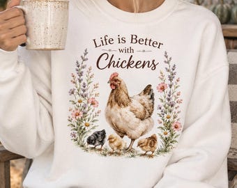 Può includere: Una felpa bianca a girocollo con una stampa grafica raffigurante una gallina marrone con tre pulcini, circondata da una ghirlanda floreale di lavanda e margherite. Il testo "Life is Better with Chickens" è stampato in marrone sopra l'immagine.