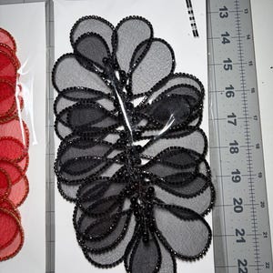 May include: Black and gray beaded applique with a floral design. The applique features a series of layered, petal-shaped elements, each outlined with small black beads. The center stem is adorned with small, round beads. The applique is on a white card.