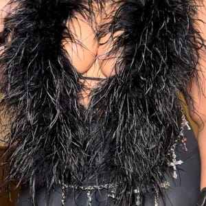Könnte beinhalten: Ein schwarzer Federboa mit einer Perlenkette und einem silbernen Anhänger. Der Boa wird über einem schwarzen Oberteil und einer schwarzen Hose getragen.