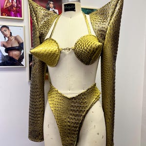 May include: A gold-colored, crocodile-embossed, three-piece outfit consisting of a bra, a pair of high-waisted bottoms, and a jacket with exaggerated shoulders.