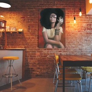 May include: A painting of a woman with an afro wearing sunglasses and a white shirt, sitting in a chair. The painting is hanging on a brick wall in a cafe setting with a wooden table and chairs.