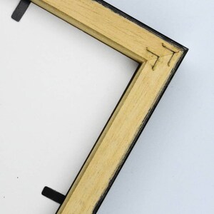 May include: Close-up of the corner of a wooden picture frame with a black painted edge. The frame is made of natural wood with a light brown colour.