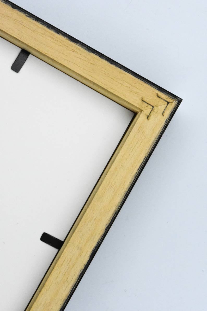 May include: Close-up of the corner of a wooden picture frame with a black painted edge. The frame is made of natural wood with a light brown colour.
