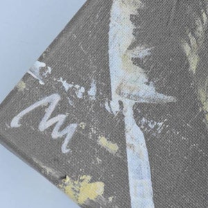 May include: Close-up of a grey canvas with white and yellow paint splatters. The canvas is signed "mw" in white paint.