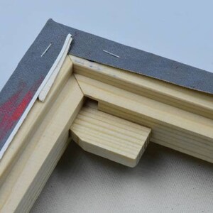 May include: Close-up of the corner of a wooden canvas frame. The canvas is stretched tightly over the frame and secured with staples. The frame is made of natural wood with a light brown finish. The corner of the frame is reinforced with a small wooden wedge.