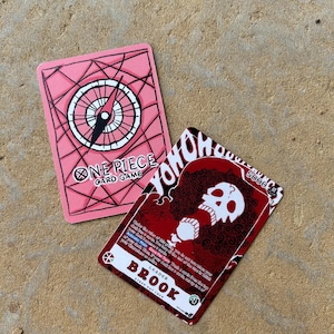 Może przedstawiać: Dwie karty do gry One Piece. Jedna karta ma różowy wzór kompasu z napisem "ONE PIECE CARD GAME". Druga karta przedstawia czerwono-białą czaszkę z napisem "BROOK" i tekstem "STRAW HAT CREW".