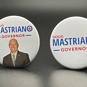 May include: Two white round buttons with a red star and blue text. The text on the buttons reads "Doug Mastriano - Governor". One button features a photo of a man in a suit.