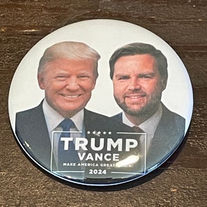 May include: A round button with a white background featuring a photo of Donald Trump and J.D. Vance. The text "TRUMP VANCE MAKE AMERICA GREAT AGAIN! 2024" is printed in black below the photo.