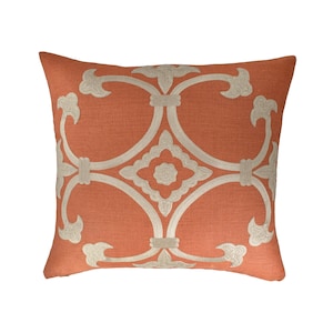 May include: An orange decorative pillow with a cream-colored embroidered geometric pattern. The pillow has a square shape and is made of a textured fabric. The embroidered design features a symmetrical, scrolling motif.