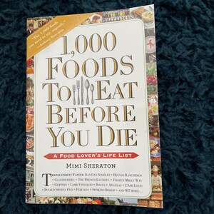 May include: A hardback book titled "1,000 Foods to Eat Before You Die" by Mimi Sheraton. The cover displays a collage of food images and text in a serif font. A quote by Anthony Bourdain is also included.