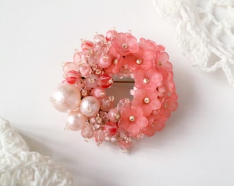 May include: A pink and white beaded flower brooch with a pearl accent. The brooch is shaped like a circle and is made of glass beads and pearls.