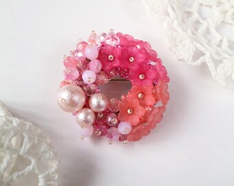 May include: A pink and white beaded flower brooch in a circular shape. The brooch is made with a variety of beads, including pearls, glass beads, and plastic flowers.