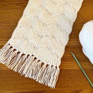 May include: A white crocheted scarf with a fringe on the bottom edge. The scarf is made with a textured stitch pattern. A ball of white yarn and a green crochet hook are also pictured.