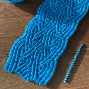 May include: A handmade, turquoise blue knitted scarf with a textured, chevron-like pattern. A ball of matching yarn and a crochet hook are also visible. The scarf is displayed on a wooden surface.