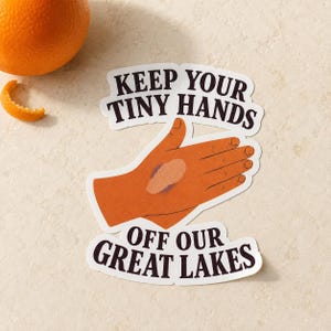 May include: A die-cut sticker featuring a graphic illustration of an orange-toned hand with a light tan palm patch. The text "KEEP YOUR TINY HANDS OFF OUR GREAT LAKES" is printed in bold, dark brown, sans-serif capital letters above and below the hand.