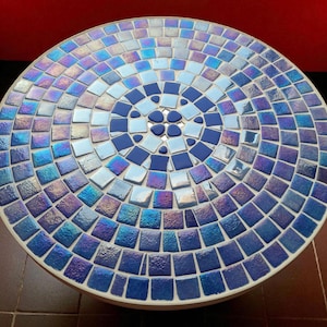 May include: Round mosaic table with a vibrant blue and iridescent tile design. The tabletop features a concentric pattern of square tiles in varying shades of blue, creating a decorative surface. The table is suitable for indoor or outdoor use.