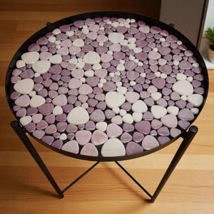May include: A round side table with a mosaic top. The mosaic is made of small, smooth, pebble-shaped tiles in shades of pink and white. The table has a black metal frame and legs. The table is approximately 24 inches in diameter.