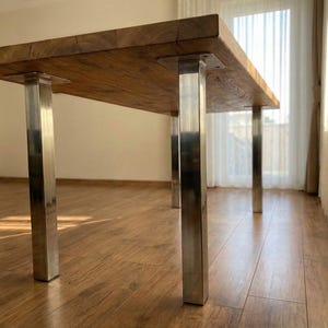 Könnte beinhalten: Ein rechteckiger Holztisch mit hellbraunem Finish. Der Tisch wird von vier Edelstahlbeinen getragen. Der Tisch steht in einem Raum mit Parkettboden und einem Fenster mit transparenten Vorhängen.