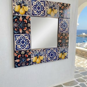 May include: Square mirror with a decorative tile frame. The tiles feature lemon and floral designs in yellow, blue, and dark blue. The mirror is mounted on a white wall, with a view of the ocean and buildings in the background.