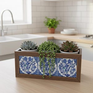 Könnte beinhalten: Ein rechteckiger Holzpflanzkasten mit drei Sukkulenten. Die Vorderseite des Kastens zeigt blau-weiße Blumentafeln. Der Kasten steht auf einer Holzoberfläche in einer Küche.
