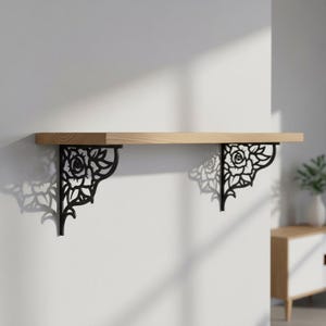 Puede incluir: Estante de madera sostenido por dos soportes metálicos negros. Los soportes presentan un diseño floral de rosas. El estante es de madera clara y está montado en una pared blanca. Los soportes miden unos 25 cm de alto.