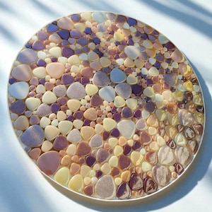 Peut inclure: Un dessous de verre rond décoratif avec un motif de mosaïque. Il est rempli de petits morceaux lisses en forme de galets dans des tons de violet, rose, beige et jaune. Le dessous de verre a une bordure blanche et est posé sur un fond blanc.