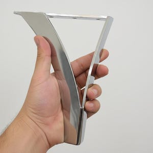 May include: A V-shaped furniture leg made of polished chrome metal with a reflective surface. The leg features a flat mounting plate at the top with three pre-drilled circular holes for attachment, and it tapers down to a narrow base.