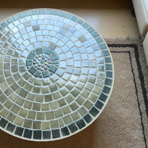 May include: Round mosaic tile table with a white border. The mosaic design features a spiral pattern of square tiles in shades of green, blue, and white. The table is on a brown rug.