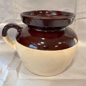 May include: A two-toned ceramic crock with a handle. The upper portion is a glossy, dark brown, while the lower half is a creamy off-white. The crock has a wide, open top and a rounded body, suggesting it's for storage or display.