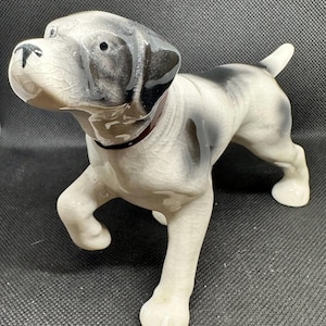 May include: A ceramic dog figurine in a walking pose. The dog has a white body with black and gray markings on its head and back. It wears a red collar. The figurine has a glossy finish and a cracked glaze effect.