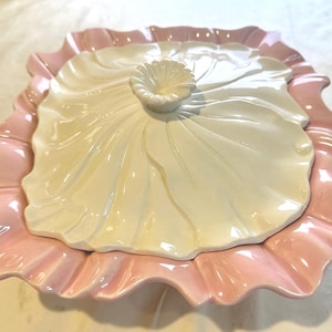 May include: A white ceramic serving dish with a pink ruffled edge. The dish has a matching lid with a flower-shaped knob.