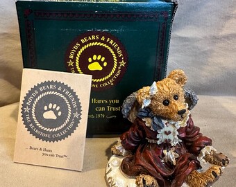 May include: A brown bear figurine wearing a red dress and a white bonnet. The bear is sitting on a white base with the words "Love is the master key" inscribed on it. The bear is part of the Boyds Bears & Friends collection.
