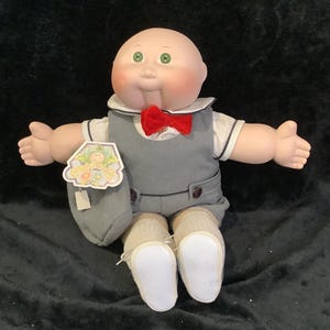 May include: A vintage Cabbage Patch Kid doll, featuring a bald head, rosy cheeks, and green eyes. The doll is dressed in a gray vest and shorts, a white shirt, a red bow tie, and white shoes. A tag is attached to the doll.