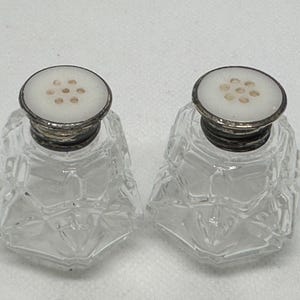 May include: A pair of clear glass salt and pepper shakers with silver tops and white porcelain lids with holes. The shakers have a faceted design.