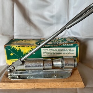 May include: Vintage nutcracker with a silver metal handle and cracking mechanism, mounted on a wooden base. The original box is visible behind the nutcracker, with text that reads "America's Best Nut Cracker."