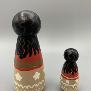 May include: Two wooden dolls with painted details. The larger doll has black hair, a red, brown, and white skirt, and white flowers. The smaller doll has the same design but is smaller in size.