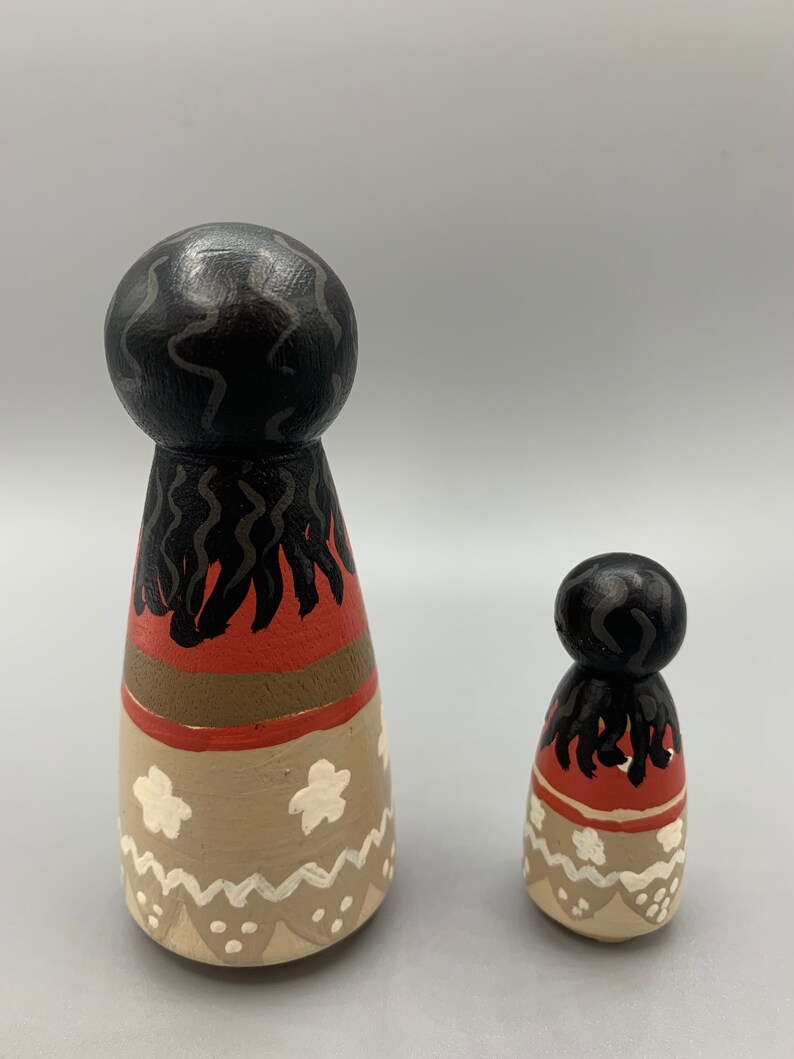 May include: Two wooden dolls with painted details. The larger doll has black hair, a red, brown, and white skirt, and white flowers. The smaller doll has the same design but is smaller in size.