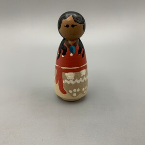 May include: A wooden doll with black hair, brown eyes, and a red and white dress. The doll is wearing a blue necklace and has white dots on its dress.