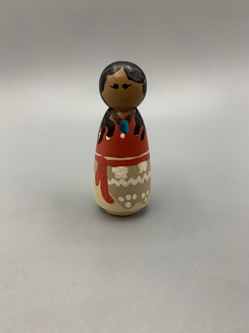 May include: A wooden doll with black hair, brown eyes, and a red and white dress. The doll is wearing a blue necklace and has white dots on its dress.