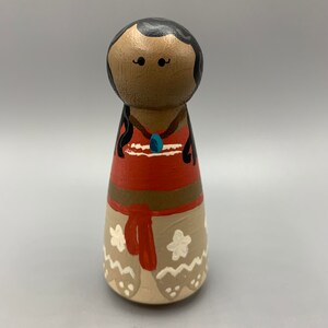 May include: A hand-painted wooden doll with brown skin, black hair, and a red and brown dress. The doll is wearing a blue necklace and has white flowers on the bottom of the dress.