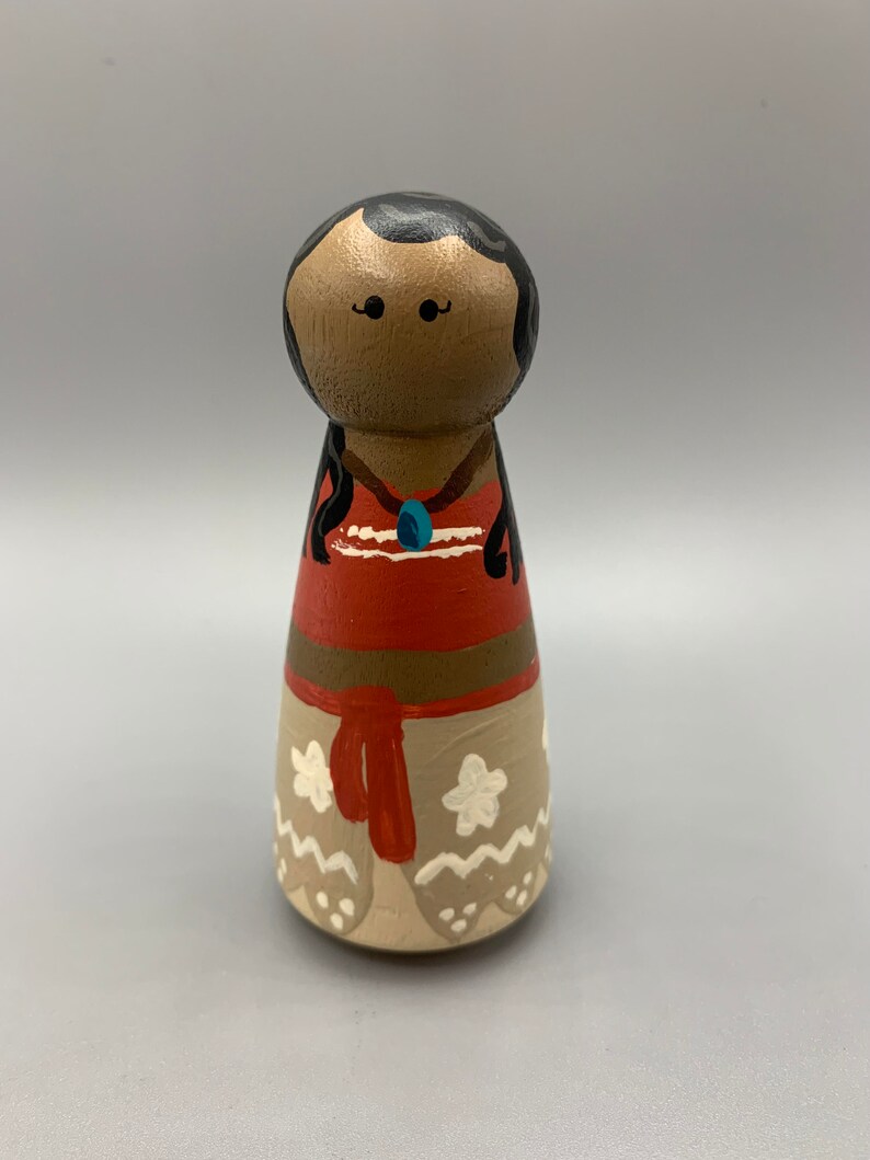 May include: A hand-painted wooden doll with brown skin, black hair, and a red and brown dress. The doll is wearing a blue necklace and has white flowers on the bottom of the dress.