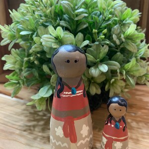 May include: Two wooden dolls, one larger than the other, are painted to resemble a mother and daughter. Both dolls have brown skin, black hair, and are wearing red and white dresses with floral patterns. The larger doll has a blue necklace.