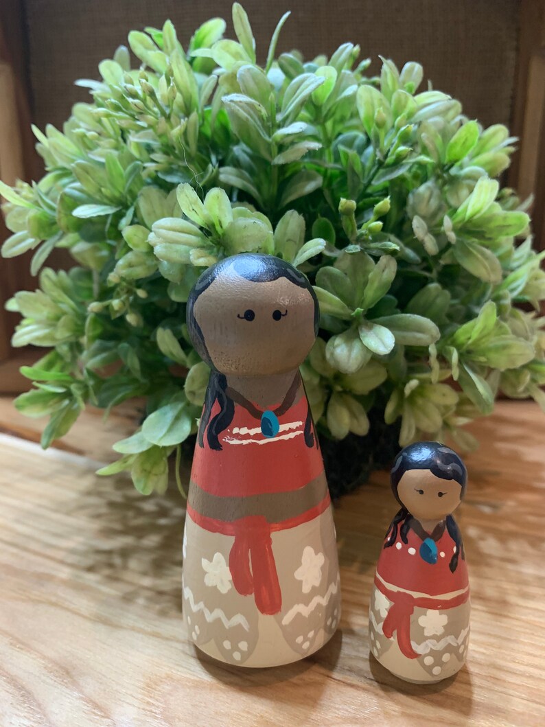 May include: Two wooden dolls, one larger than the other, are painted to resemble a mother and daughter. Both dolls have brown skin, black hair, and are wearing red and white dresses with floral patterns. The larger doll has a blue necklace.