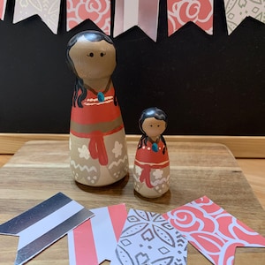 May include: Two wooden dolls with painted faces and hair. The larger doll is wearing a red and brown dress with white accents. The smaller doll is wearing a red dress with white accents. Both dolls have black hair and brown eyes. The dolls are sitting on a wooden surface with pink and white patterned paper flags.