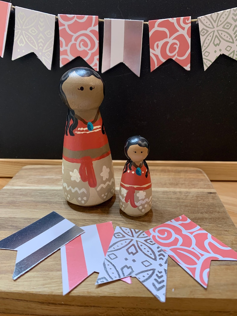 May include: Two wooden dolls with painted faces and hair. The larger doll is wearing a red and brown dress with white accents. The smaller doll is wearing a red dress with white accents. Both dolls have black hair and brown eyes. The dolls are sitting on a wooden surface with pink and white patterned paper flags.