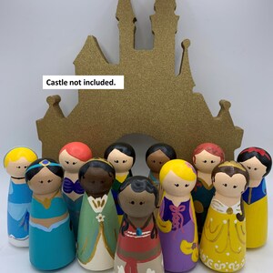 May include: A set of 11 hand-painted wooden peg dolls dressed as Disney princesses. The dolls are arranged in front of a gold castle cutout. Each doll is sold separately. "Castle not included." and "Each doll sold separately." are written in the image.