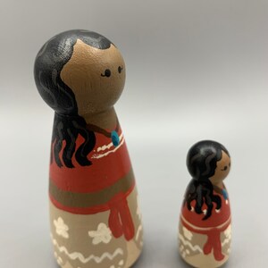 May include: Two wooden dolls with painted faces and black hair. The larger doll is wearing a red and brown dress with a white and brown skirt. The smaller doll is wearing a similar dress.
