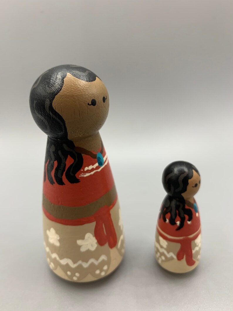 May include: Two wooden dolls with painted faces and black hair. The larger doll is wearing a red and brown dress with a white and brown skirt. The smaller doll is wearing a similar dress.