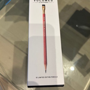 Peut inclure: Une boîte blanche avec le texte "VOLUMES by BLACKWING" et un crayon rouge. Le crayon a une virole et une gomme dorées. La boîte indique également "12 LIMITED EDITION PENCILS".