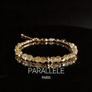 Op de afbeelding: Een gouden armband met gele edelsteen kralen. De armband wordt weerspiegeld op een zwart oppervlak. De tekst "PARALLELE PARIS" wordt eronder in het wit weergegeven.